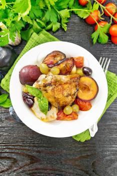 Baked chicken with tomatoes, apples, plums and grapes in a plate on a napkin, garlic, parsley and basil on dark wooden board background from above