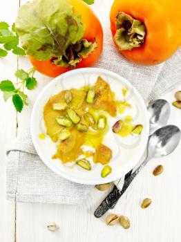 Dessert of yogurt, persimmon and honey with vanilla, cardamom and pistachios in a bowl on a napkin, mint and spoon on the background of light wooden board on top