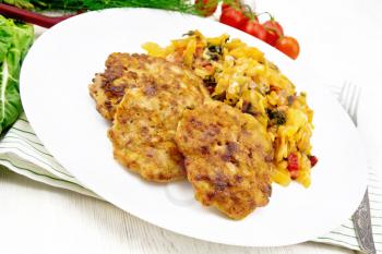 Fritters of minced meat with stewed cabbage in a plate, fork on kitchen towel, tomatoes, parsley and chard on light wooden board background
