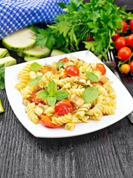 Fusilli with chicken, zucchini and tomatoes in a plate, napkin, fork, basil and parsley on black wooden board background
