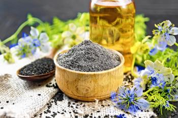 Flour of black caraway in a bowl, seeds in a spoon on burlap, oil in bottle and twigs Nigella sativa with blue flowers and leaves on wooden board background