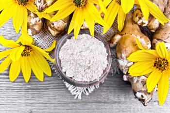 Jerusalem artichoke flour in a clay bowl on a burlap with flowers and vegetables on wooden board background from above