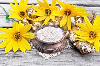 Jerusalem artichoke flour in a clay bowl on a burlap with yellow flowers and vegetables on wooden board background