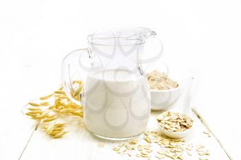 Oat milk in a jug, flour in bowl, oatmeal in spoon, stalks oaten on the background of light wooden board