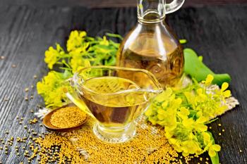 Mustard oil in a glass gravy boat and decanter, grains in a spoon, leaves and yellow mustard flowers on burlap against dark wooden board