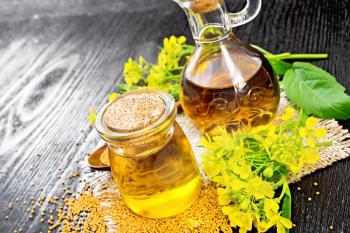 Mustard oil in a glass jar and decanter, mustard seeds on burlap, flowers and leaves on black wooden board background