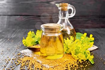 Mustard oil in a glass jar and decanter, mustard grains on a burlap napkin, flowers and leaves on wooden board background
