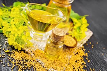 Mustard oil in two glass jars and a gravy boat, grains, leaves and yellow mustard flowers on burlap on wooden board background