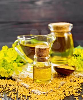 Mustard oil in two glass jars and a gravy boat, grains, leaves and yellow mustard flowers on a burlap napkin on wooden board background