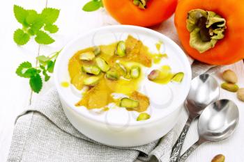 Dessert of yogurt, persimmon and honey with vanilla, cardamom and pistachios in a bowl on a napkin, mint and spoon on background of light wooden board