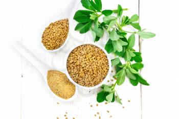 Fenugreek seeds in a bowl and spoon, ground spice in a spoon with green leaves on wooden board background from above