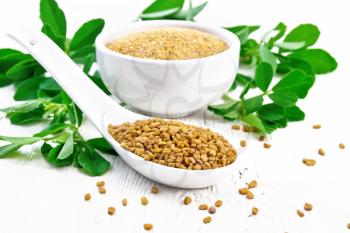 Fenugreek seeds in a spoon and ground spice in a bowl with green leaves on background of light wooden board