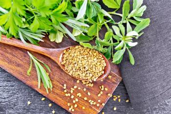 Fenugreek seeds in a spoon on a brown plate with spicy herbs, a dark napkin on wooden board background from above