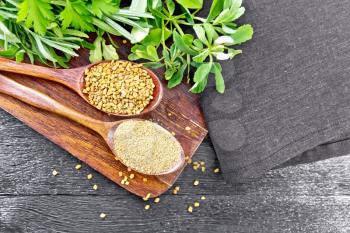 Fenugreek seeds and ground spice in two spoons on brown plate with herbs, a napkin on wooden board background from above