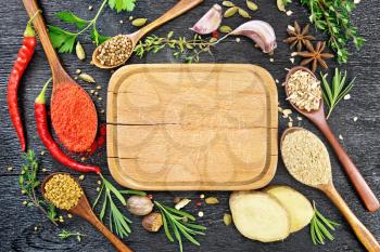 Frame of plate, spoons with ground pepper, fenugreek, ginger, coriander, sprigs of fresh rosemary, thyme and parsley, pods of hot pepper, cardamom, anise, nutmeg and garlic on black wooden board top background