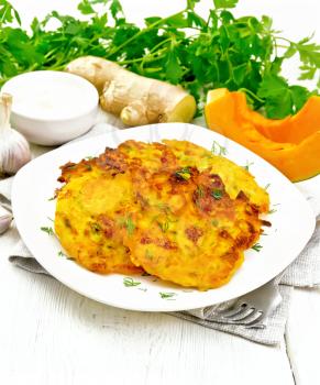 Pumpkin fritters in a plate on napkin, sour cream in a bowl, parsley, garlic and ginger on background of light wooden board