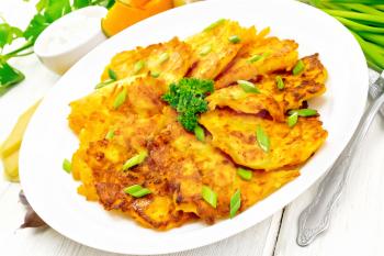 Pumpkin and cheese fritters with ginger in a white plate, sour cream in a bowl, napkin, parsley, garlic and fork on background of light wooden board