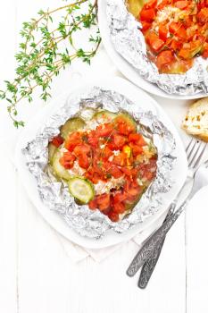 Pink salmon with zucchini, tomatoes, onions, garlic and thyme, baked in foil on a plate, napkin, fork and bread on the background of a light wooden board from above