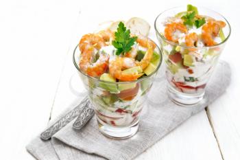 Puff salad with shrimp, avocado, fresh cucumber, sweet pepper and tomato, seasoned with yogurt sauce in two glass glasses on a napkin, bread and forks on a light wooden board background
