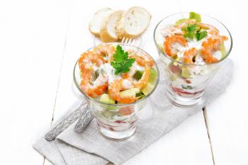 Puff salad with shrimp, avocado, fresh cucumber, sweet pepper and tomato, seasoned with yogurt sauce in two glass glasses on a towel, bread and forks on a white wooden board background
