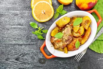 Turkey stewed with peaches, fresh hot pepper and orange sauce, basil leaves in a roasting pan, napkin, fruits on black wooden board background from above