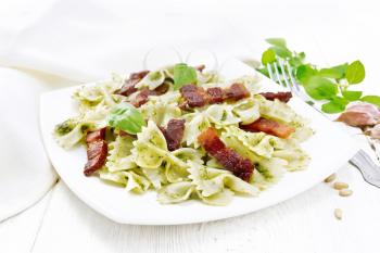 Farfalle pasta with pesto sauce, fried bacon and basil in a plate, garlic, fork and napkin on white wooden board background