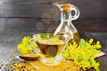 Mustard oil in a glass gravy boat and decanter, grains in a spoon, leaves and yellow mustard flowers on burlap on wooden board background