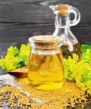 Mustard oil in a glass jar and decanter, mustard seeds on burlap, flowers and leaves on wooden board background