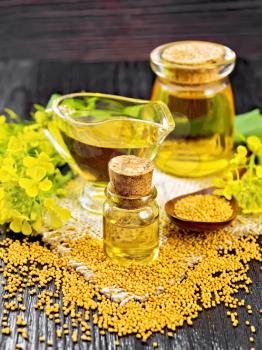 Mustard oil in two glass jars and a gravy boat, grains, leaves and yellow mustard flowers on burlap on a dark wooden board background
