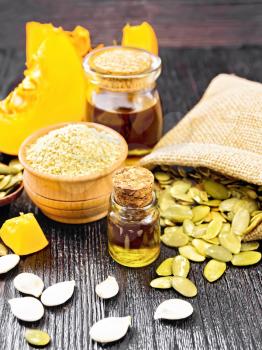 Pumpkin oil in glass vial and a jar, flour in a bowl, seeds in a spoon and bag, slices of vegetable on dark wooden board background