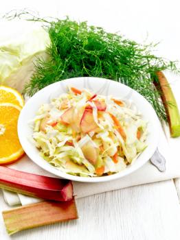 Fresh cabbage, carrot and rhubarb salad with orange juice, honey and mayonnaise dressing in a plate on a towel, dill and fork on white wooden board background
