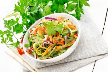 Spicy salad of cucumbers, carrots, chili peppers, purple onions, cilantro and black sesame seeds, seasoned with vinegar and lemon juice in a bowl on a napkin on wooden board background