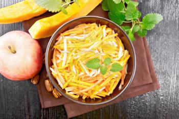 Pumpkin, carrot and apple salad with pecans seasoned with vegetable oil in a bowl on a napkin, mint on dark wooden board background from above