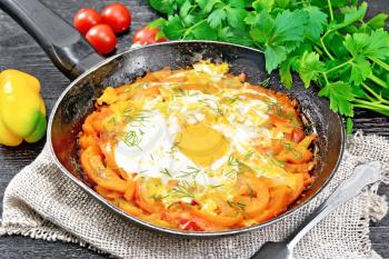 Fried eggs with tomatoes, sweet pepper, onions and herbs in a pan on burlap, parsley, fork on black wooden board background