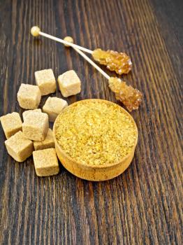 Granulated brown sugar in a bowl, crystal on a stick and sugar cubes on dark wooden board background