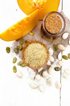 Pumpkin flour in a bowl and oil in a jar on burlap, seeds on table and slices of vegetable on wooden board background from above