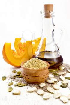 Pumpkin flour in a bowl and oil in a decanter on burlap, seeds on table and slices of vegetable on background of light wooden board