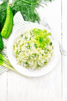 Salad of fresh cabbage, green onions and cucumber with vinegar and vegetable oil dressing in a plate, towel, dill and fork on wooden board background from above