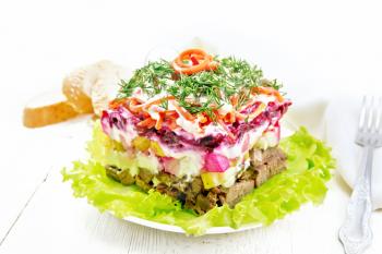 Puff salad with beef, potatoes and beets, pears, Korean spicy carrots, seasoned with mayonnaise and garnished with dill on a green lettuce in a plate, towel, bread on light wooden board background