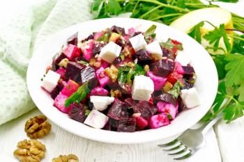 Salad with beets, salted feta cheese, apples, walnuts, parsley, seasoned with balsamic vinegar and olive oil in a plate, a towel and a fork on a wooden board background