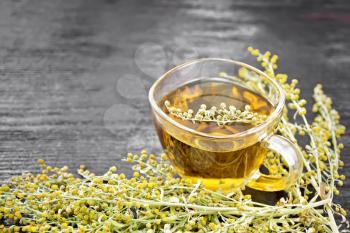 Gray wormwood herbal tea in a glass cup, fresh sagebrush flowers on dark wooden board background