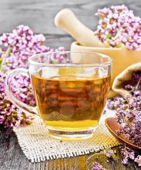 Oregano herbal tea in a glass cup on burlap, fresh flowers in mortar and on the table, dried marjoram flowers in a bag and spoon on wooden board background