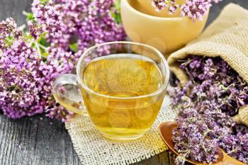 Oregano herbal tea in a glass cup on burlap napkin, dried marjoram flowers in a bag and spoon, fresh flowers in mortar and on table on wooden board background