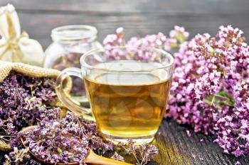 Oregano herbal tea in a glass cup, fresh flowers, dried marjoram flowers in a bag, jar and spoon on wooden board background