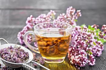 Oregano herbal tea in a glass cup, fresh flowers and metal strainer with dried marjoram flowers on dark wooden board
