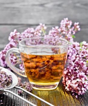 Oregano herbal tea in a glass cup, fresh flowers and metal strainer with dried marjoram flowers on wooden board
