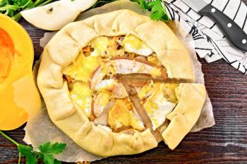 Pie of pumpkin, pear, soft cheese and walnuts on parchment, napkin and knife, parsley on a wooden board background from above