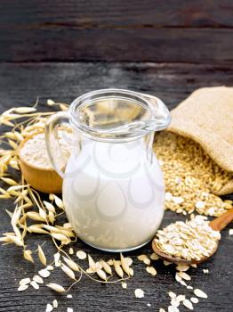 Oat milk in a jug, flour in bowl, oatmeal in a spoon, grain in bag, oaten stalks on a wooden board background