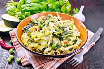 Tagliatelle pasta with zucchini, green peas, asparagus beans, hot peppers and spinach in a plate on towel, garlic, fork and basil on the background of dark wooden board