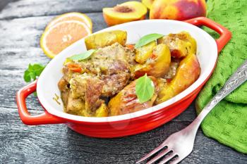 Turkey stewed with peaches, fresh hot pepper and orange sauce, basil leaves in a roasting pan, napkin, fruits on black wooden board background